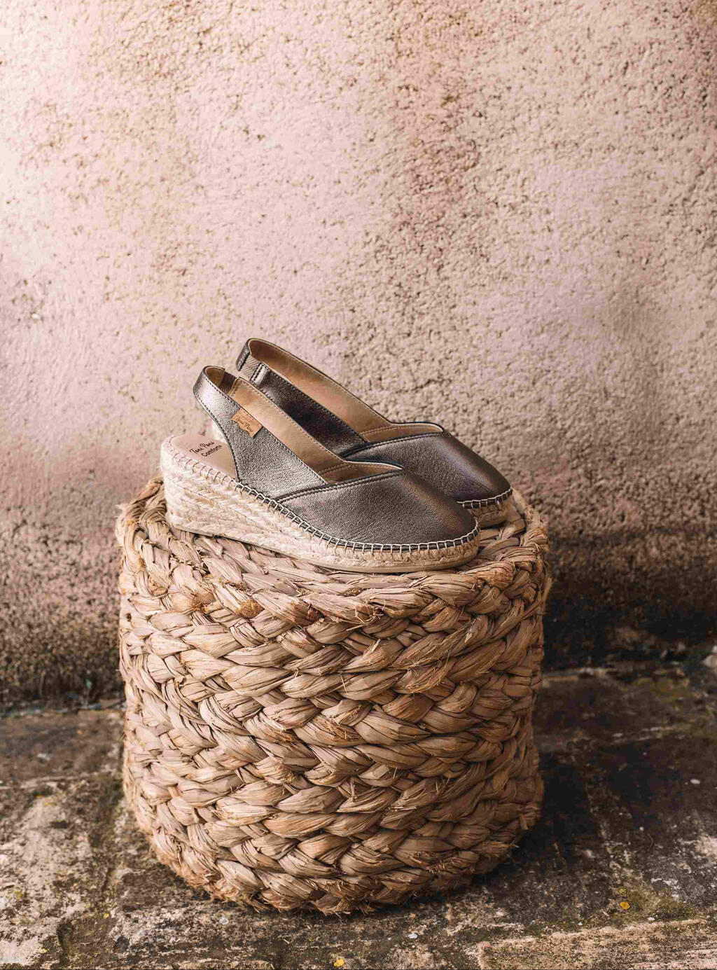 Pair of silver wedge sandals on a woven stool against a textured wall.